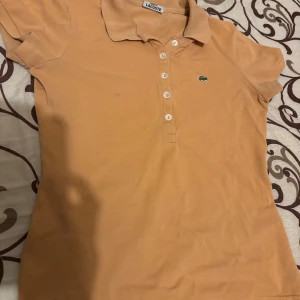 Beige pikétröja från Lacoste - Säljer en klassisk beige pikétröja från Lacoste med korta ärmar och krage. Tröjan har knappar framtill och den ikoniska gröna krokodilloggan på bröstet. Perfekt för en avslappnad och stilren look.