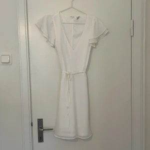 Vit klänning med volangärmar - Elegant vit klänning med v-ringning och volangärmar. Den har ett knytband i midjan för en smickrande passform. Perfekt för sommarens tillställningar.