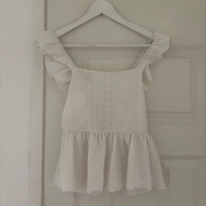 Vit blus med volang och knyt - Supersöt vit blus med volangdetaljer på axelbanden och broderade hålmönster. Blusen har en luftig, peplum-liknande nederdel och knytning i ryggen för extra charm. Perfekt för dig som gillar romantisk och feminin stil.