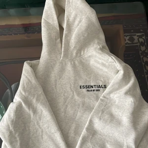 Ljusgrå Essentials Fear of God hoodie -  Har inte använt en gång. Säljer en ljusgrå hoodie från Fear of God Essentials med svart logotyptryck både fram och bak. Kommer med originalpåse. Kan byta mot hoodies helst burberry eller denim