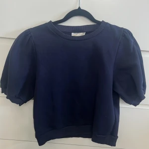 Marinblå puffärm tröja Gina Tricot - Snygg marinblå tröja från Gina Tricot med puffiga korta ärmar och rund hals. Tröjan har ribbade muddar vid hals, ärmslut och nederkant. Mjuk och skön i bomullsmaterial, perfekt för dig som gillar stilrena och trendiga detaljer.