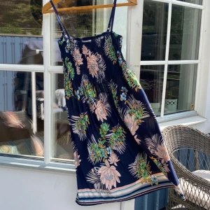 Blommig marinblå klänning Lucky Style - Säljer en marinblå klänning från Lucky Style i storlek M/L med tunna axelband och blommigt mönster i ljusa toner. Klänningen är tillverkad av 65% viskos och 25% polyester, har en avslappnad passform och når till knäna. Perfekt för sommarens varma dagar!