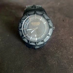 Svart klocka från Versus Versace - Snygg svart klocka från Versus Versace med rund urtavla och diskret mönstrad bakgrund. Klockan har tydliga index, datumvisning och svart armband med Versus-logga och lejonhuvud. Perfekt accessoar för dig som gillar stilrena detaljer.