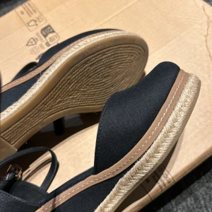 Svarta sandaler från Tommy Hilfiger - Snygga svarta sandaler från Tommy Hilfiger med rund tå och rem runt ankeln. Ovandelen är i textil och sulan har en snygg detalj i repimitation. Perfekta för sommaren med diskret logga på sidan och bekväm platt sula.