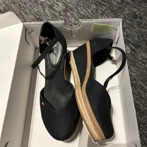 Svarta espadrille sandaler Tommy Hilfiger - Snygga svarta espadrille sandaler från Tommy Hilfiger med kilklack och justerbar rem runt ankeln. Ovandel i textil, sula med flätad detalj och liten logga på sidan. Perfekta för sommarens alla outfits.