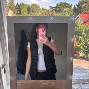 Svart dunväst med dragkedja - Säljer en svart, quiltad dunväst med hög krage och dragkedja framtill. Västen har en klassisk, enkel design och är perfekt att slänga över en hoodie eller t-shirt. Passformen är normal och materialet är lätt och smidigt.