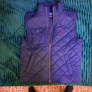 Quiltad blå dunväst från Polo Ralph Lauren - Snygg quiltad blå dunväst från Polo Ralph Lauren med klassisk röd logga broderad på bröstet. Västen har hög krage, dragkedja framtill, en bröstficka med knapp och två sidofickor. Perfekt för lager-på-lager och stilren look.