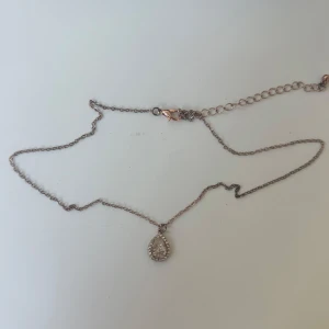 Rosé halsband med droppformad berlock - Snyggt och stilrent halsband i rosé med tunn kedja och en droppformad berlock. Berlocken är dekorerad med glittriga stenar runt en större mittsten som ger extra bling. Perfekt för dig som gillar diskreta men ändå iögonfallande smycken.
