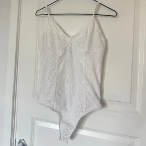 Vit spetsig korsettbody med smala band - Säljer en vit korsettinspirerad body med smala axelband och v-ringning. Hela bodyn är täckt av vacker spets med blommigt mönster och har en figurnära passform. Perfekt att styla med jeans eller kjol för en trendig look. (Obs fick den med en liten fläck)