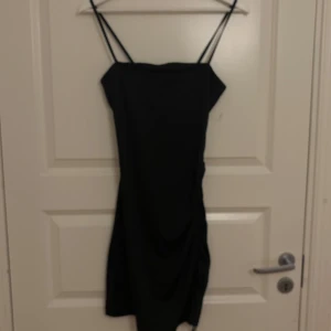 Svart omlottklänning från H&M Divided - Snygg svart kortklänning från H&M Divided med smala axelband och omlottdetalj på sidan. Klänningen har en tight passform och är tillverkad i mjuk polyester som sitter skönt mot kroppen. Perfekt för dig som gillar minimalistisk stil och vill ha en enkel men trendig look.