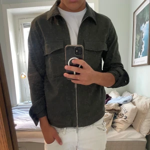 Grön overshirt från Zara med dragkedja - Säljer en stilren grön overshirt från Zara med dragkedja framtill och två stora bröstfickor. Jackan är i bomull och har klassisk krage och långa ärmar. Perfekt att slänga på över en t-shirt för en clean look.