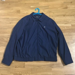 Blå vindjacka från Polo Ralph Lauren XL - Snygg marinblå vindjacka från Polo Ralph Lauren. Storlek XL men passar mer som L. Jackan har dragkedja framtill, klassisk krage och broderad logga på bröstet. Tillverkad i polyester och har elastisk midja för en schysst passform. Perfekt för dig som gillar stilren och sportig look.