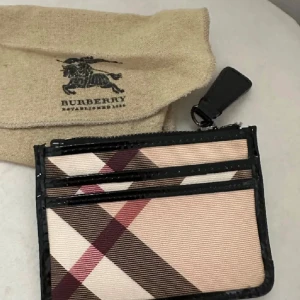 Burberry plånbok med dragkedja - Snygg plånbok från Burberry i klassiskt rutig design med beige, bruna och röda toner. Plånboken har svarta detaljer, flera kortfack och dragkedja upptill. Kommer med originalpåse