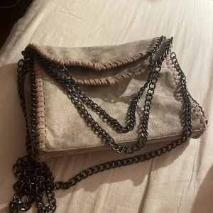 Beige axelväska med kedja och mockadetaljer - Snygg beige axelväska med grova svarta kedjor och dekorativa sömmar längs kanterna. Väskan har ett lock och dragkedjefack på baksidan. Materialet är mockaliknande och insidan har flera fack för småprylar. Perfekt för dig som gillar en cool och trendig look.