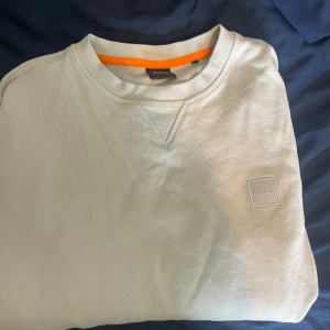 Ljusgrå sweatshirt från BOSS - Säljer en stilren ljusgrå sweatshirt från BOSS i storlek S. Tröjan har rund halsringning, diskret BOSS-logga på bröstet och en orange detalj på insidan av kragen. Perfekt för en clean och avslappnad look.