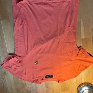Orange t-shirt från Lyle & Scott - Snygg orange t-shirt från Lyle & Scott med klassisk gul broderad logga på bröstet. T-shirten har rund halsringning och korta ärmar. Perfekt för dig som gillar stilrena plagg med en färgklick. Materialet är mjuk bomull som känns skönt mot huden.