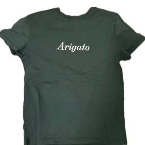 Grön Arigato t-shirt i bomull - Cool grön t-shirt från Arigato med vit text på bröstet. Klassisk rund hals och korta ärmar, perfekt för en chill och stilren look. Tillverkad i mjuk bomull som känns skön mot huden. Passar dig som gillar minimalistisk design.