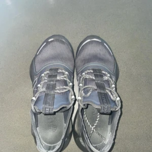 Svarta Adidas sneakers med vita detaljer - Snygga svarta Adidas sneakers med klassiska vita ränder på sidorna och robust svart sula. Ovandelen är i mesh och syntet, med coola reflekterande detaljer och snörning framtill. Perfekta för dig som gillar sportig och modern stil.