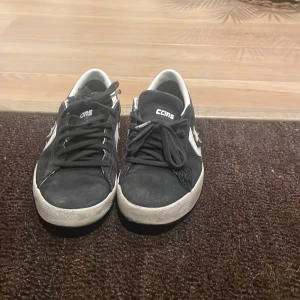 Svarta Converse CONS sneakers i mocka - Svarta Converse CONS sneakers med vit sula och vita detaljer, inklusive stjärnlogga på sidan och bak. Skorna har snörning och är tillverkade i mocka med inslag av syntet. Klassisk låg modell med rund tå och platt sula, perfekt för en streetwear-look.