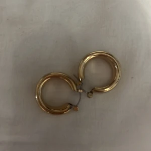 Guldiga chunky hoops örhängen - Säljer ett par snygga chunky hoops i guldton. De är runda och har en klassisk, stilren design som passar till många olika looks. Perfekta för dig som gillar statement-smycken och vill addera lite extra bling till din outfit.