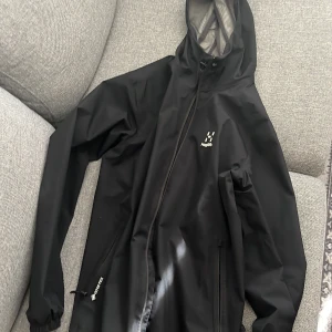 Svart regnjacka från Haglöfs med huva - Svart regnjacka från Haglöfs med huva och dragkedja framtill. Jackan har minimalistisk design, elastiska muddar och två fickor med dragkedja. Materialet är syntet och jackan är perfekt för blöta dagar. Klassisk logga på bröstet och justerbar nederkant. Jackan är i bra skick och har storlek L