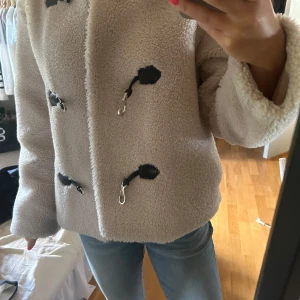 Beige teddyjacka med coola detaljer - Säljer en beige teddyjacka med snygga svarta spänndetaljer framtill. Jackan är fluffig och har lång ärm, perfekt för dig som gillar en mysig men trendig look. Passformen är normal och materialet är mjukt och värmande. Perfekt till jeans och sneakers.