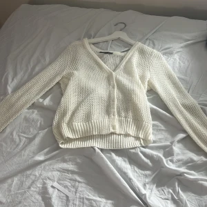 Vit stickad kofta med v-ringning - Säljer en vit stickad kofta med v-ringning och knappar framtill. Koftan har ett luftigt, grovstickat mönster och ribbade muddar vid ärmslut och nederkant. Perfekt att slänga över en t-shirt eller linne för en chill look.