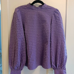 Lila blus med puffärm och struktur - Säljer en lila blus med snyggt rutigt strukturmönster och puffiga långärmar. Blusen har hög hals och breda muddar vid ärmsluten som ger en cool siluett. Perfekt för dig som gillar att sticka ut med färg och form!
