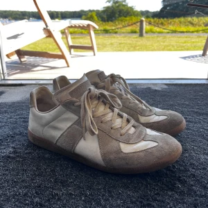 Maison Margiela skor - Maison Margiela Replica sneakers i beige och vitt med klassisk retrodesign. Skorna har ovandel i skinn och mocka, snörning och bruna gummisulor. Perfekta för dig som gillar minimalistisk och stilren vibe. Saknar alla tillbehör då skorna är gamla. Storlek 44, bra skick förutom sulan vilket syns på sista bilden 7/10 men äkta, pris går att diskutera✅
