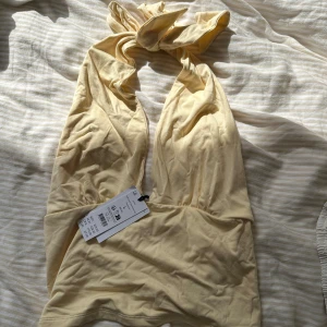 Gul halterneck linnetopp från Monki - Supersöt ljusgul halterneck topp med knyt i nacken och djup v-ringning. Toppen är gjord i mjuk bomull, perfekt för varma dagar. Lapparna kvar 