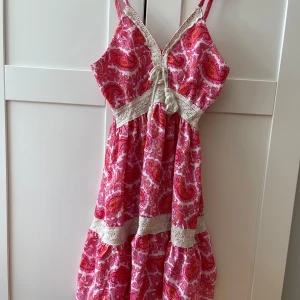 Rosa paisley sommarklänning med spets - Säljer en superfin kortklänning med paisleymönster i rosa, rött och orange toner. Klänningen har spetsdetaljer i beige, snörning framtill och smala axelband. Materialet känns lätt och luftigt, perfekt för varma dagar. Volangkant nedtill ger extra flow.
