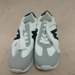 Gråvita sneakers med svart detalj - Snygga sneakers i vitt och ljusgrått med svart blixtformad detalj på sidan. Skorna har rund tå, platt sula och klassisk snörning. Perfekta för dig som gillar stilren och sportig look. Materialet är en mix av syntet och textil.