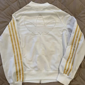 Adidas kofta  - Snygg vit Adidas bomberjacka med klassiska guldiga tre ränder längs ärmarna och broderad logga på bröstet. Jackan har dragkedja framtill, ribbade muddar och två fickor. 