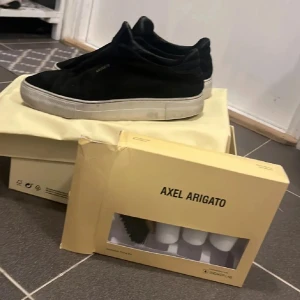 Svarta sneakers från Axel Arigato - Säljer ett par svarta sneakers från Axel Arigato med vit platt sula. Skorna har en stilren design utan snörning och är tillverkade i mocka. Perfekta för dig som gillar minimalistisk stil.