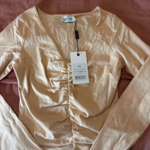 Beige omlottopp från Ivy Revel XS - Snygg beige omlottopp från Ivy Revel i storlek XS. Toppen har lång ärm, v-ringning och cool rynkdetalj framtill som ger en trendig look. Tillverkad i mjuk bomull, perfekt för dig som gillar stilrena och moderna plagg.