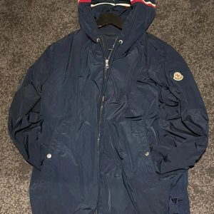 Marinblå vindjacka från Moncler - Snygg marinblå vindjacka från Moncler med huva och dragsko. Jackan har dragkedja framtill, två fickor med tryckknappar och klassisk Moncler-logga på ärmen.  Toppskick. Storlek M 