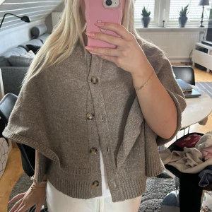 Beige kofta med knappar och cape-look - Säljer en beige kofta med cool cape-design och stora knappar framtill. Tröjan är croppad och har ribbade kanter. Perfekt för dig som vill sticka ut med en unik och trendig stil.