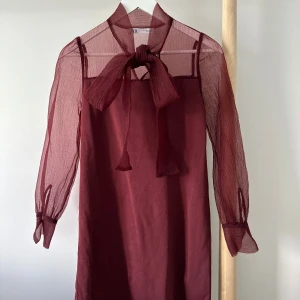Vinröd kortklänning med rosett från Zara - Vinröd kortklänning från Zara med transparenta långärmar och stor knytrosett i halsen. Klänningen har en lätt A-linjeform och är gjord i ett tunt, syntetiskt material som ger en luftig känsla. 