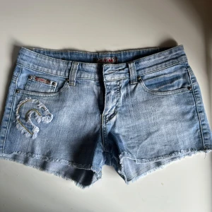 Ljusblå jeansshorts från C.T.M - Snygga ljusblå jeansshorts från C.T.M med fransig kant och klassisk femficksdesign. Shortsen har en broderad och sliten C-detalj på ena fickan och coola slitningar för en avslappnad vibe. Perfekta för varma dagar och chill häng.