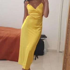 Gul långklänning med smala axelband - Säljer en snygg gul långklänning med smala axelband och omlott-detalj vid bysten. Klänningen har en figurnära passform och är tillverkad i ett glansigt material som ger en lyxig känsla. Perfekt för dig som vill sticka ut med färg och stil.