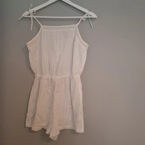 Vit somrig byxdress från H&M - Superfin vit byxdress från H&M med tunna axelband som knyts och spetsdetaljer längs kanten. Toppen har diskreta ränder och små prickar i tyget, samt resår i midjan för en snygg passform. Perfekt för varma dagar och chill vibes. Har tyvärr vuxit ur denna, men den har inga tydliga täckande på användning.