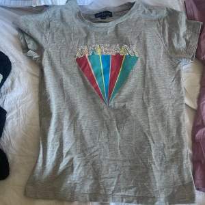 Grå t-shirt med paljetter från XOXO friends - Grå t-shirt från XOXO friends med korta ärmar. Framsidan har ett stort diamantmotiv i blått, rött och gult samt texten 'DREAM' i glittriga paljetter. Skön och mjuk bomullskänsla, perfekt för dig som gillar färgglada detaljer.