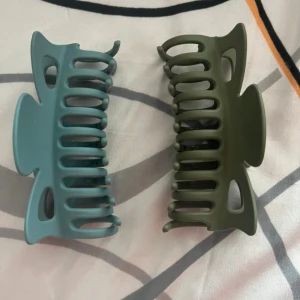 Grön och blå hårklämma i plast - Två stora hårklämmor i matta färger, en i dämpad blå och en i olivgrön. Båda har en klassisk rektangulär form med breda grepp och öppna sidor, perfekta för att hålla upp håret snyggt och enkelt. Stilrena och passar till alla outfits.