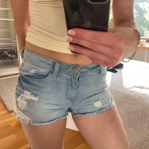 Ljusa jeansshorts med spetsdetaljer - Säljer ett par ljusblå jeansshorts med råa kanter, slitningar och snygga vita spetsdetaljer på sidorna. Modellen är lågmidjad och har klassiska fem fickor. Perfekta för sommarens alla häng! Passar xs