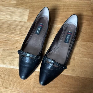 Svarta ballerinaskor från Ecco i skinn - Vintage stil, klassiska svarta ballerinaskor från Ecco i skinn med spetsig tå och låg klack. Snygg detalj med rem och knapp framtill samt kontrastsömmar. 