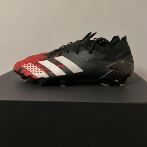 Adidas Predator fotbollsskor svart/röd - Snygga Adidas Predator fotbollsskor i svart och rött med vita ränder. Skorna har snörning, mönstrad ovandel och dobbar för bästa grepp på planen. Tillverkade i syntetmaterial med coola detaljer och en aggressiv look som sticker ut på matchen. Saknar inte heller några taggar på skorna.