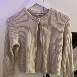Beige ribbad kofta från 157, stl S - Superfin beige ribbad kofta från 157 i storlek S. Koftan har rund halsringning, långa ärmar och knäppning framtill med små vita knappar. Perfekt att slänga över en topp för en chill och stilren look.