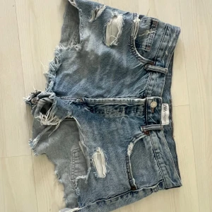 Blå slitna jeansshorts från Zara - Riktigt snygga blå jeansshorts från Zara med hög midja och rejält slitna detaljer. Shortsen har fransiga kanter, flera hål och en cool, avslappnad vibe. Perfekta för sommaren och festivaler. Klassisk femficksmodell med bälteshällor.