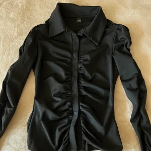 Svart skjorta med rynkade detaljer - Stilren svart skjorta från Shein med långa ärmar och snygga rynkade detaljer längs knappraden. Skjortan har klassisk krage och är figurnära i modellen, perfekt för dig som gillar en trendig och elegant look.