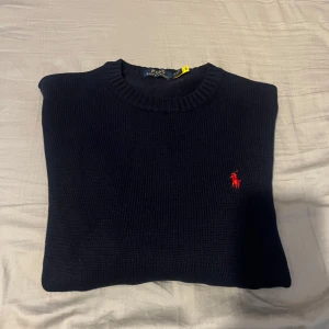 Ralph Lauren tröja  - Snygg mörkblå stickad tröja från Polo Ralph Lauren i storlek M. Klassisk rund hals och långärmad modell med den ikoniska röda Polo-loggan broderad på bröstet. Perfekt för dig som gillar stilrena och tidlösa plagg med premiumkänsla.